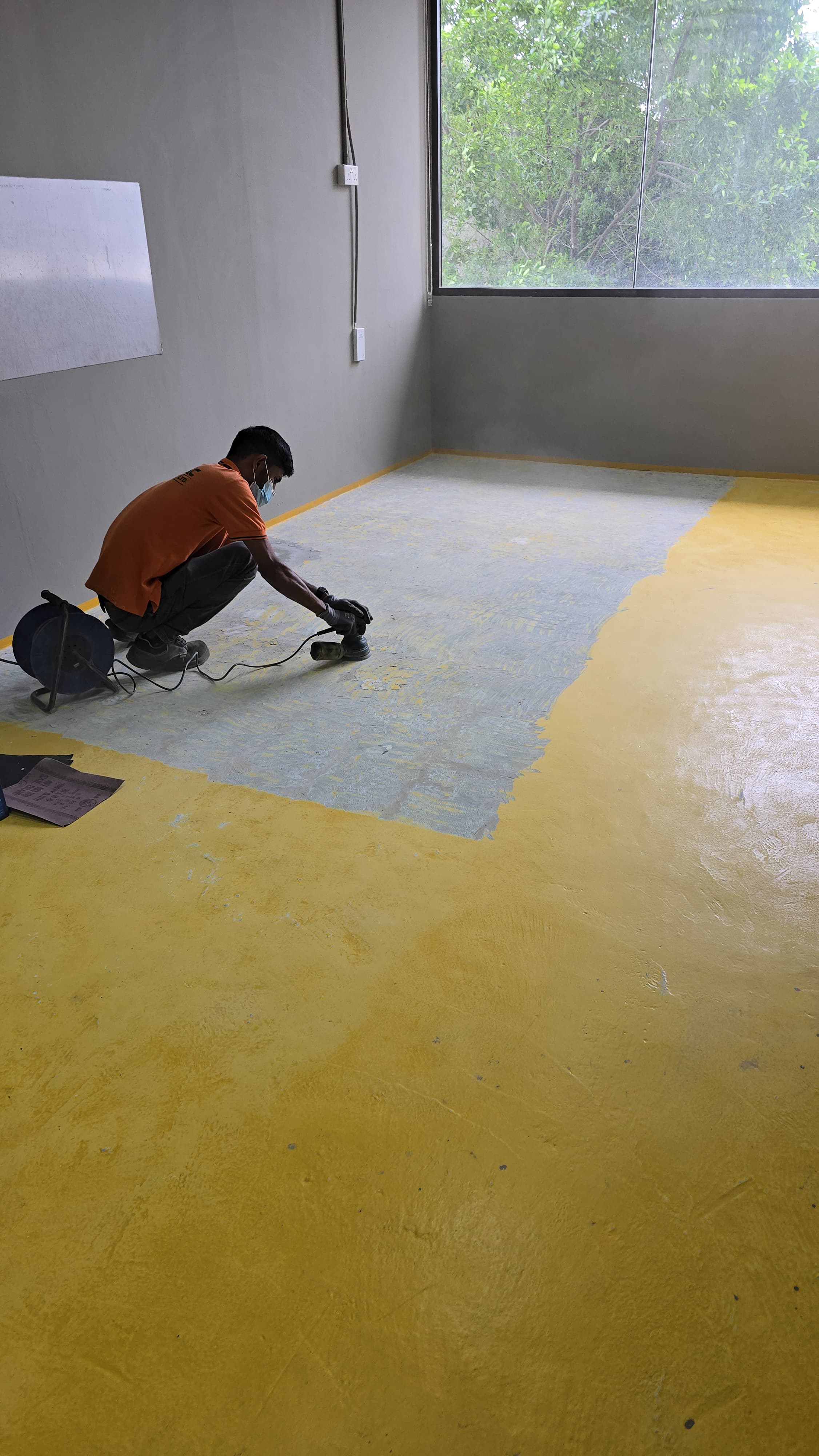 A worker in an orange shirt sands a bright yellow wall with a power tool.
