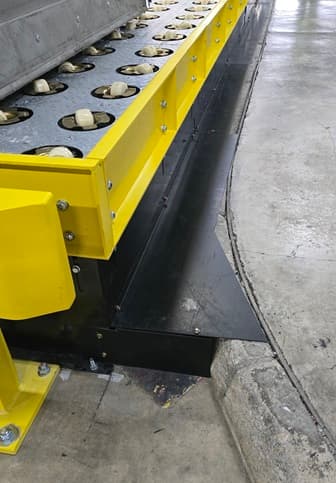 Industrial conveyor featuring white omnidirectional rollers and bright yellow safety guards on a concrete floor.