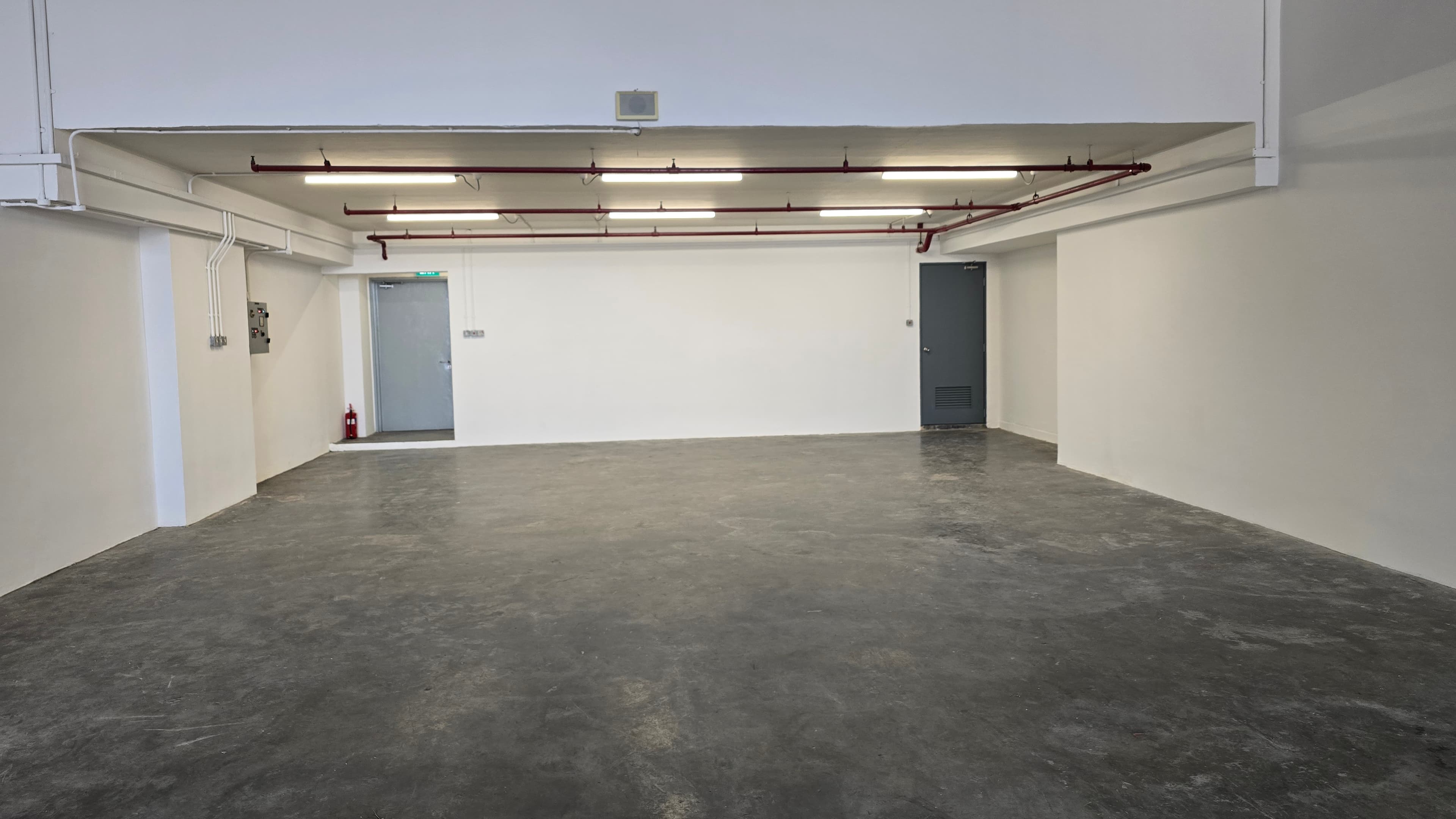 Empty industrial space with concrete floors, white walls, red overhead pipes, and two doors.