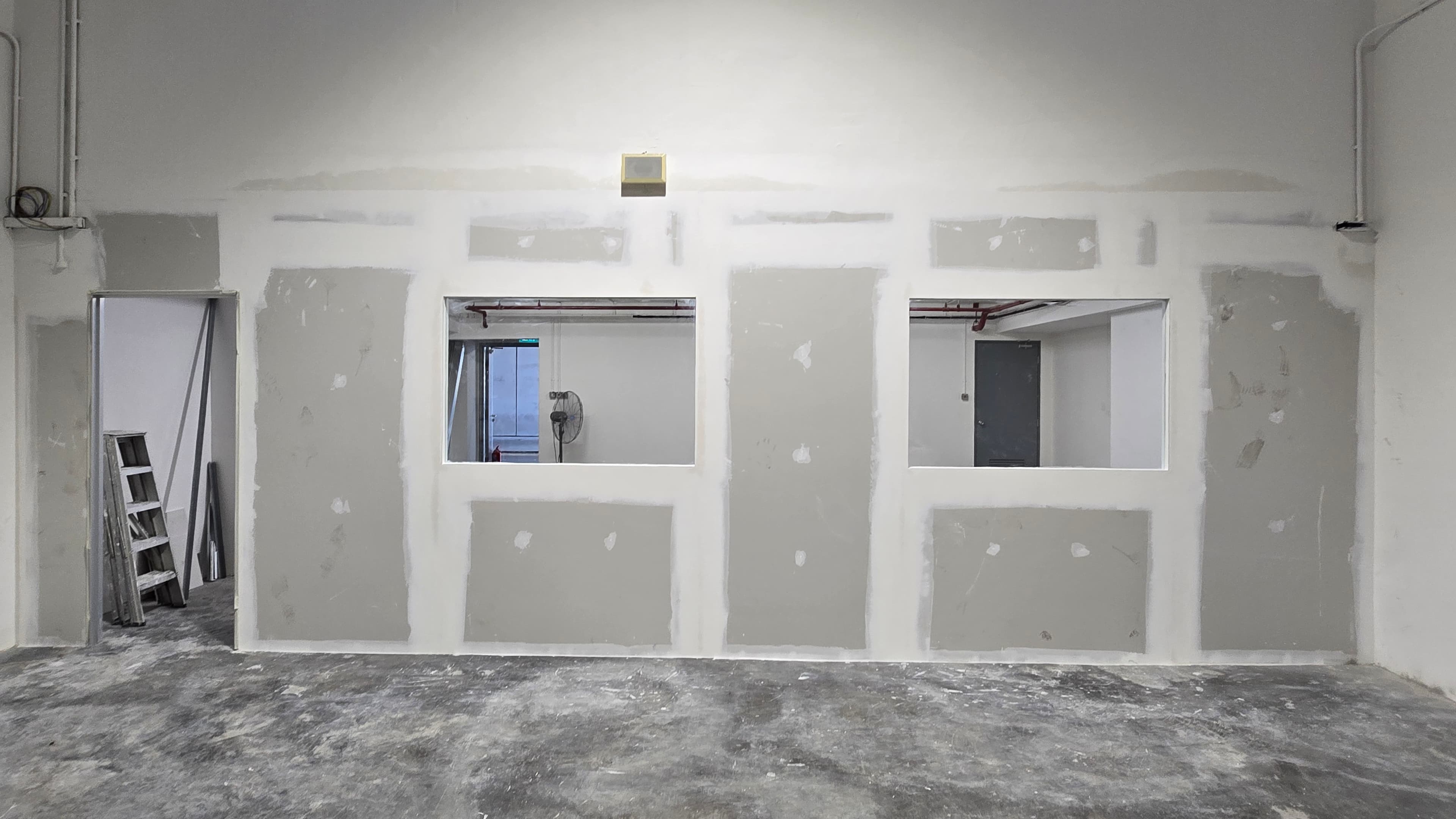 Unfinished drywall partition with two window openings and a doorway in a dusty construction area.