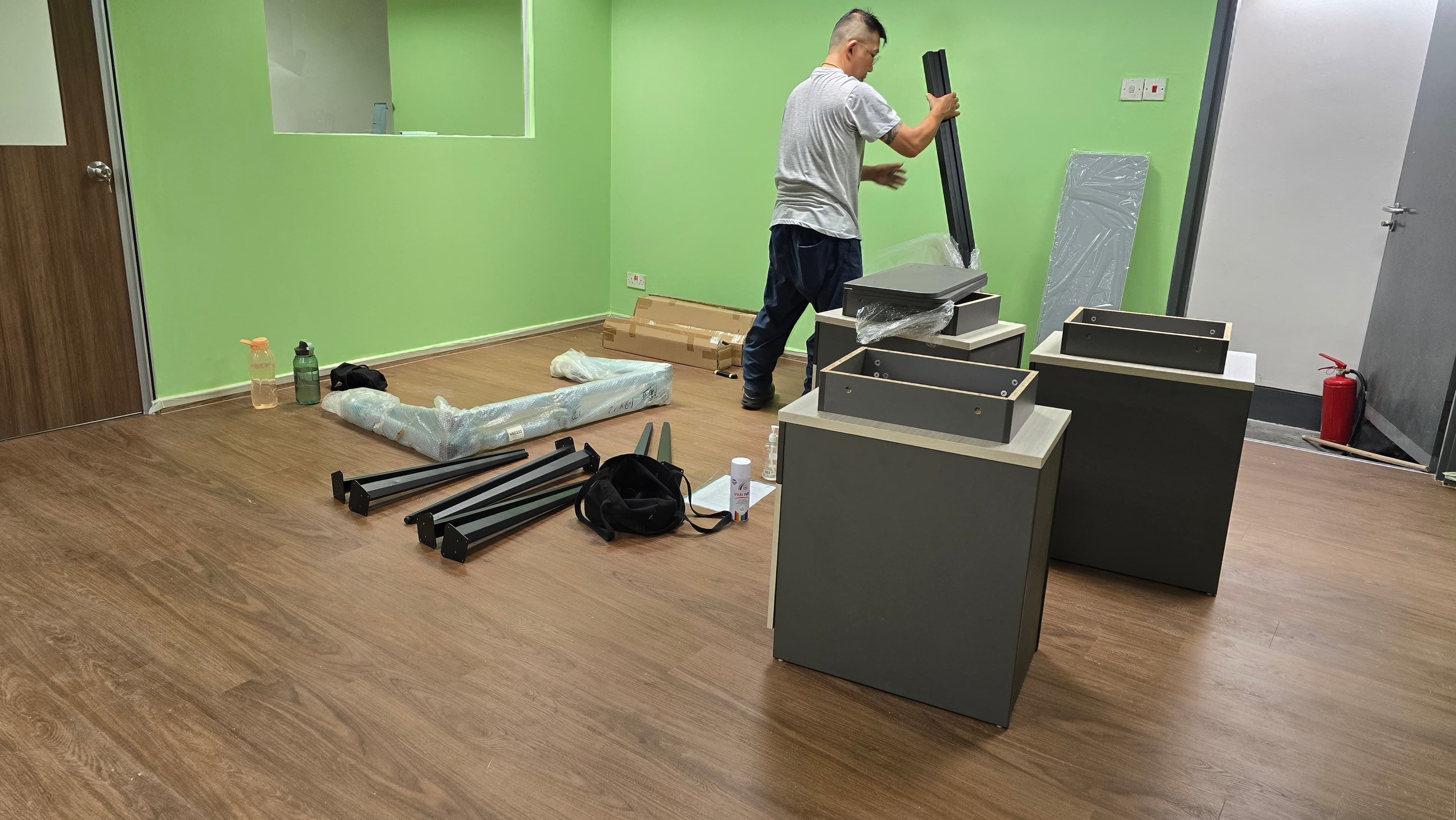 Man assembling grey office furniture in a room with green walls and wooden floors.