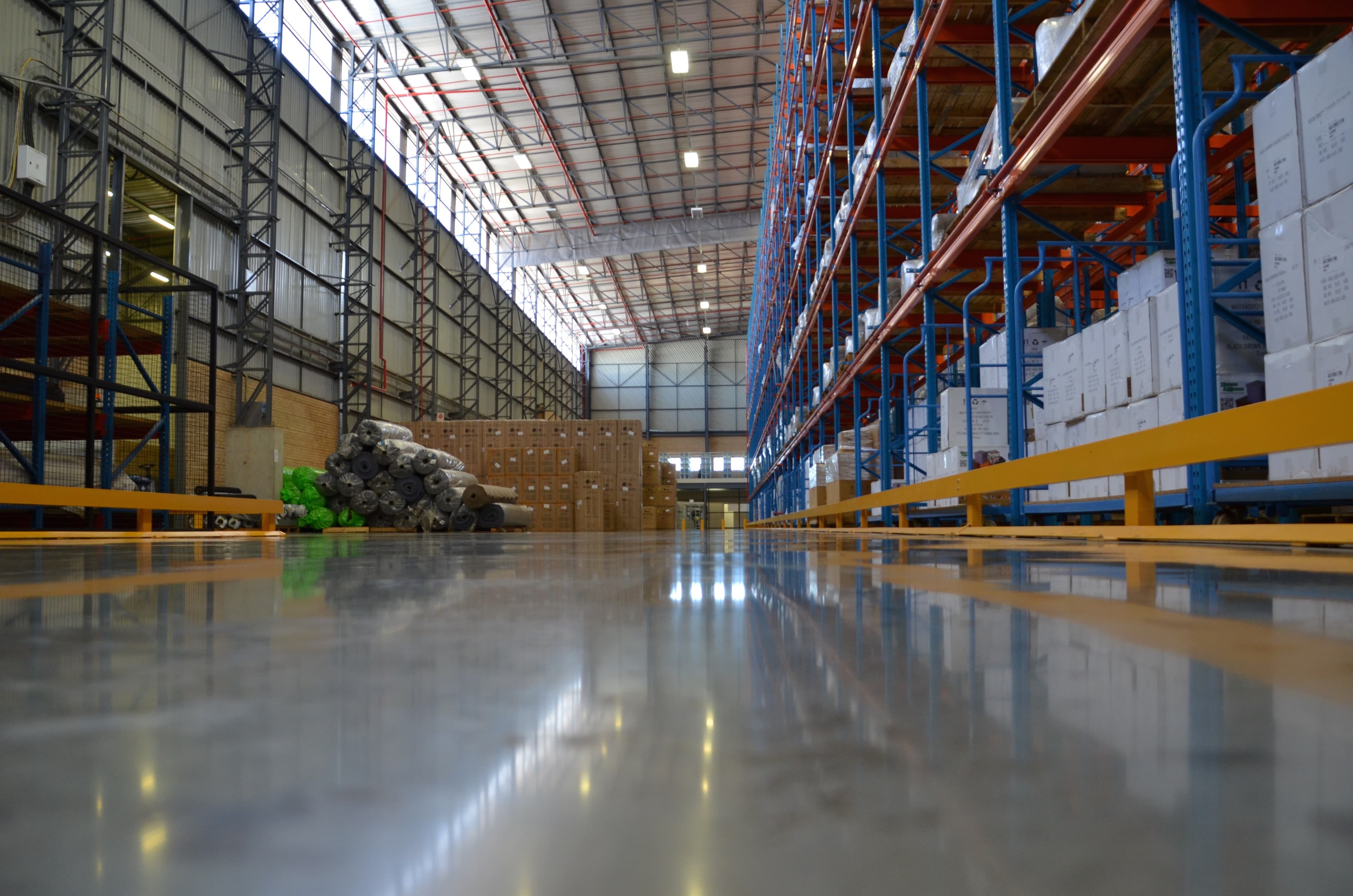Polished warehouse floor reflecting tall blue storage racks filled with boxes and stacked industrial rolls.