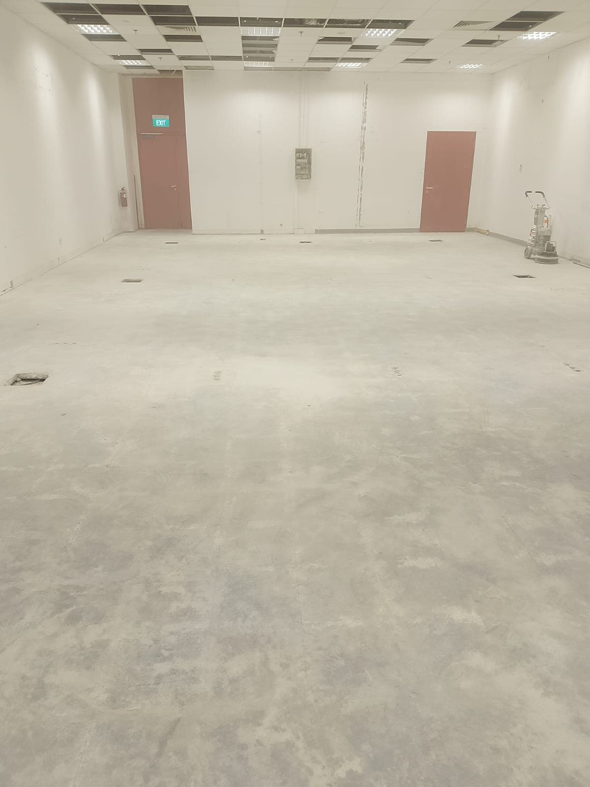 Large empty room under renovation with concrete floors, two red doors, and a floor grinder.