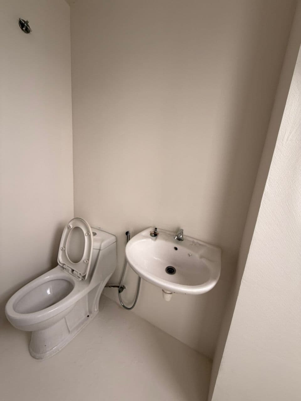 White toilet with open lid and a small wall-mounted sink against plain beige walls.