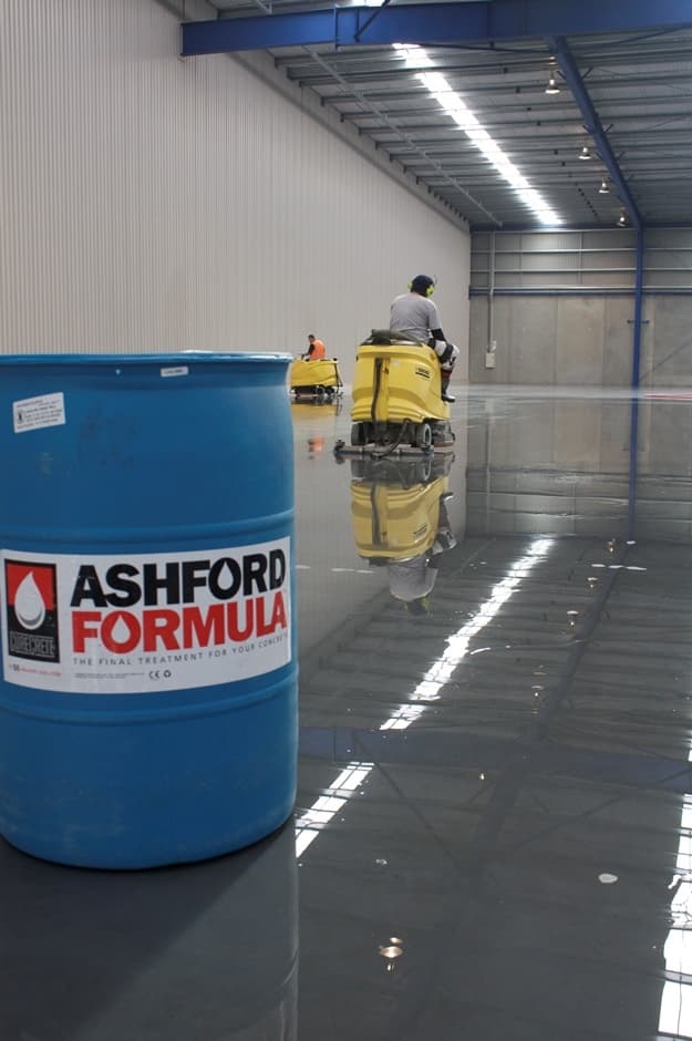 Blue Ashford Formula drum with workers cleaning a shiny warehouse floor using yellow industrial scrubbers.