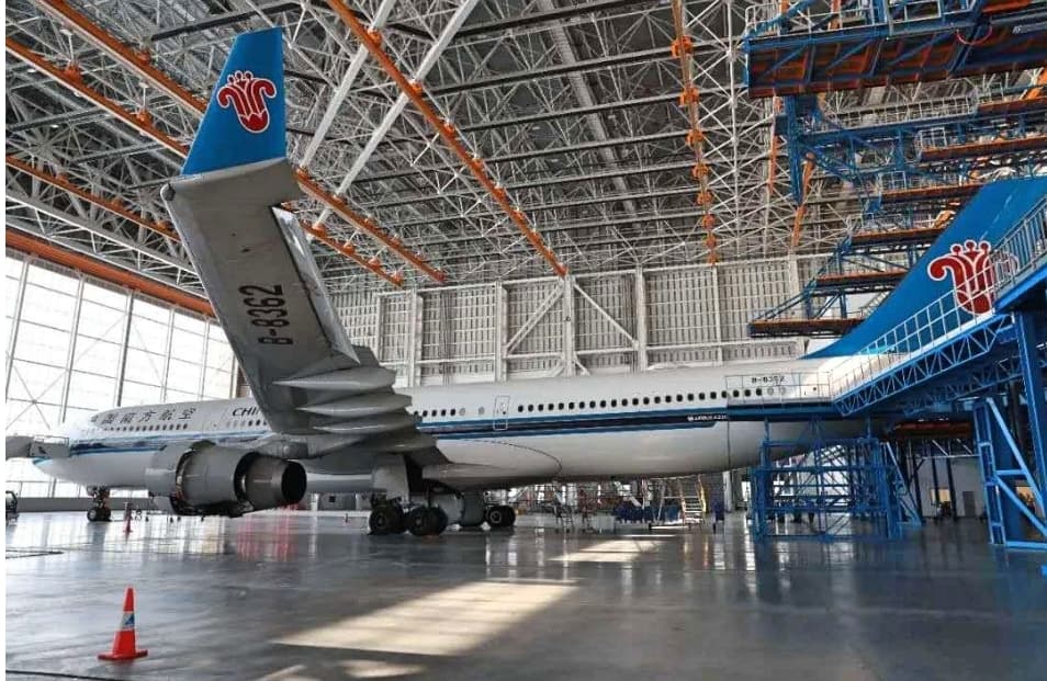 China Southern Airlines Airbus A330 parked inside a large maintenance hangar with blue metal scaffolding.