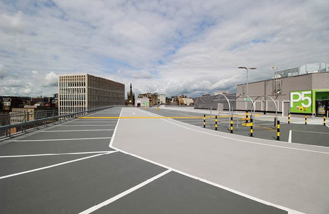 Empty rooftop parking lot with white markings and city buildings under a cloudy sky.