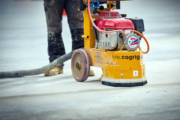 Worker using a yellow CoGri floor grinding machine with a vacuum hose on concrete.