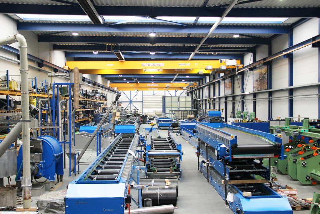 Large industrial workshop featuring blue conveyor belts and yellow overhead cranes for manufacturing operations.